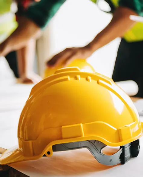 A Yellow Hardhat on a Wooden Table with Some Constrution Workers in the Background Putting Their Fists Together in Agreement