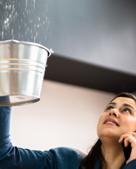 Water Damage Restoration Contractors in Jamaica, NY - A Woman on the Phone Catching Water from a Ceiling Leak with a Silver Bucket