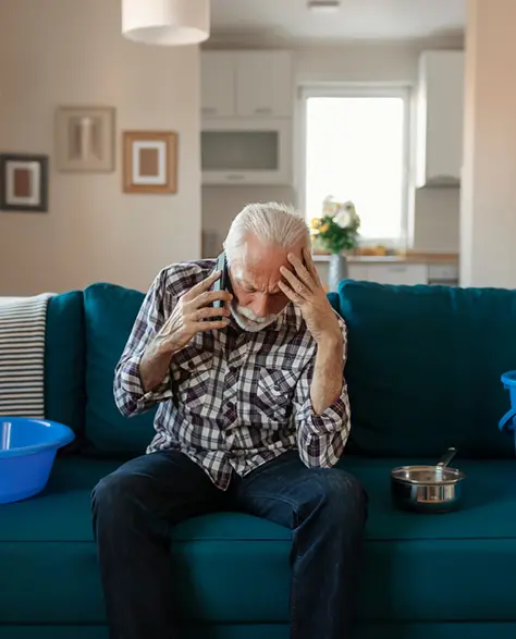 Water Damage Restoration Contractors in Glen Cove, NY - An Elderly Man Sitting on the Couch Talking on the Phone with Buckets Next to Him on the Couch to Catch a Roof Leak