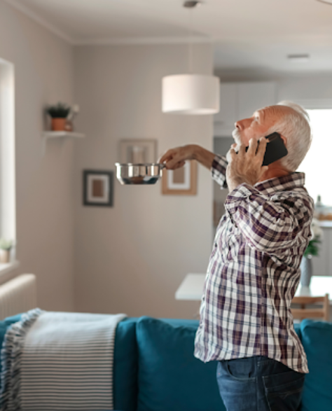 Water Damage Restoration Contractors in Borough Park, NY -  An old man looking distraught is holding a steel pan under to catch water leaks from his roof in the living room while on his mobile