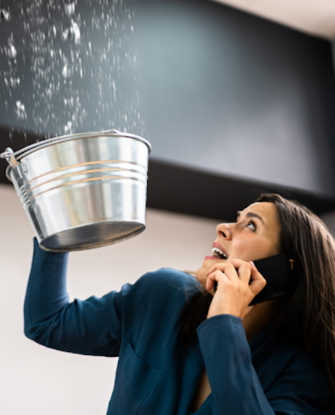 Water Damage Restoration Contractors in Corona, NY - A woman holding a pail to collect water from a leaking roof in one hand and holding a phone in her other hand. 