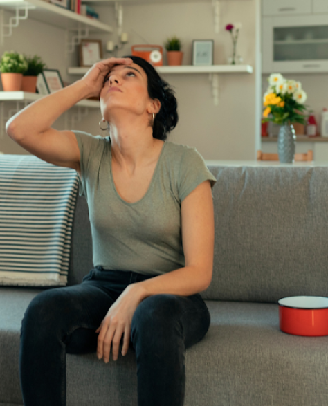  Water Damage Restoration Contractors in Flushing, NY: A Woman with Her Hand On Her Forehead Looking at the Ceiling with a Pot Sitting Next to Her On the Couch to Catch the Water from a Leak<br />
