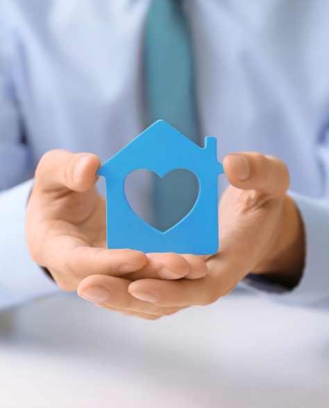 Water Damage Restoration Contractors in Kew Gardens, NY - A Woman Sitting at the Table Holding a Blue House with a Heart Cut Out of the Middle Made of Paper<br />
