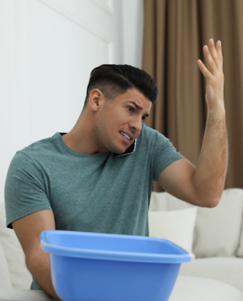 Water Damage Restoration Contractors in Rego Park, NY - A Man with a Bucket to Catch Water from a Leak Sitting on His Couch Talking on the Phone<br />
