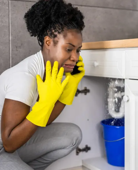 Water Damage Restoration Contractors in Holtsville, NY - A Woman with Gloves on Calling Someone to Help After Placing a Bucket Under Her Sink