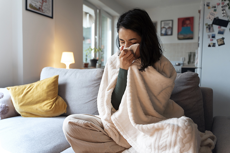 A woman sitting cross legged on a sofa with a draped blanket and cleaning her nose