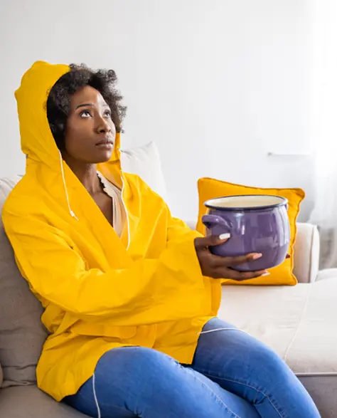 Water Damage Restoration Contractors in Richmond Hill, NY - A Woman Sitting on Her Couch With a Bucket to Catch Rainwater From a Leak in the Roof Wearing a Rain Coat