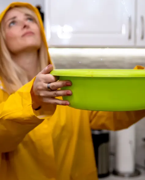 Water Damage Restoration Contractors in South Ozone Park, NY - A Woman with a Yellow Raincoat On Holding a Bucket Under a Roof Leak