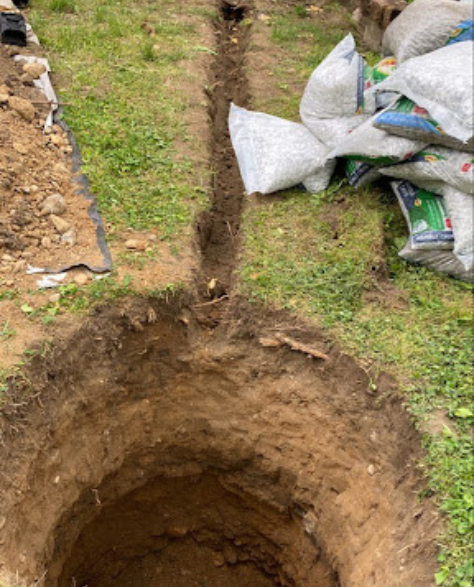 Foundation Repair Contractors in Valley Stream, NY - The Beginning of a French Drain Installation Showing a Hole Dug in the Ground and a Trench Dug from it to the House<br />
