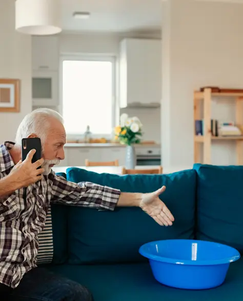 Water Damage Restoration Contractors in Woodside, NY - A Man Complaining On the Phone Pointing at a Bucket on the Couch that is Catching Water from a Roof Leak