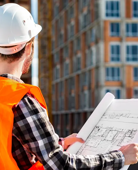 A contractor in an orange hard hat holding a blueprint in his hand and looking at a building 