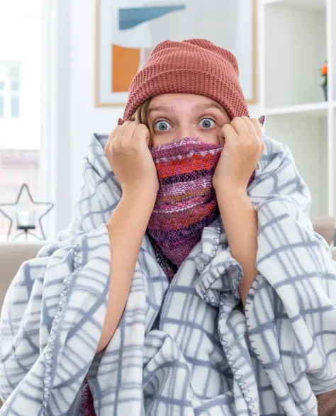 A man wrapped in blanket and beanie sitting in cold drafts in his home which is not air sealed