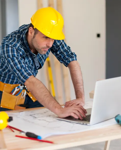 A contractor in a hard hat leaning on his study table working on his laptop<br />
