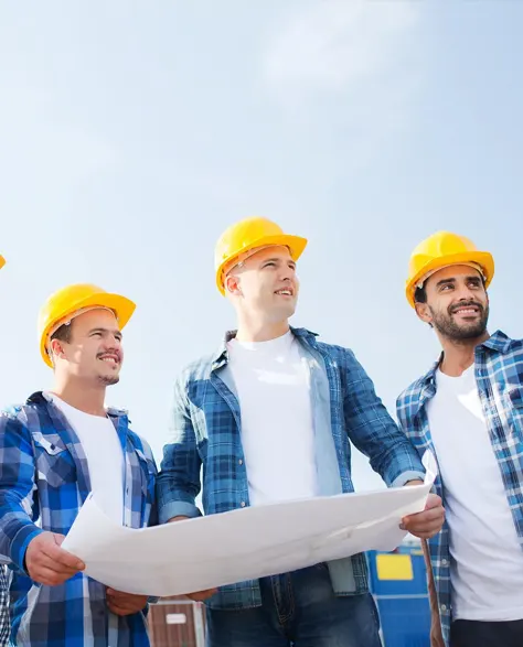 Contractors in yellow hard hats studying a concrete structure while holding a blueprint and standing at a construction site