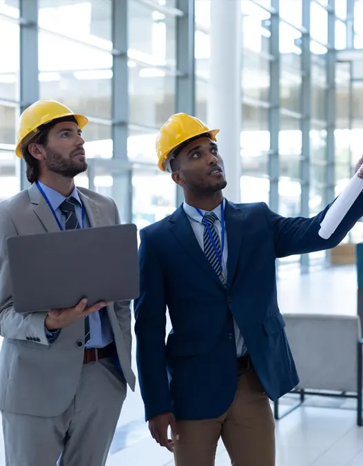 Two contractors in hard hats standing in a building observing a distant blue print.