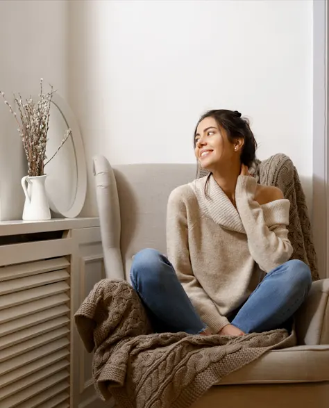 A Woman sitting and smiling on a sofa in a cozy environment in her home near a heater