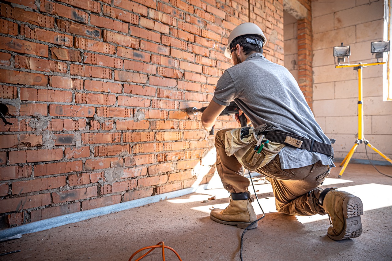 Construction worker in safety helmet and work boots using a power drill on a brick wall inside a building under renovation.