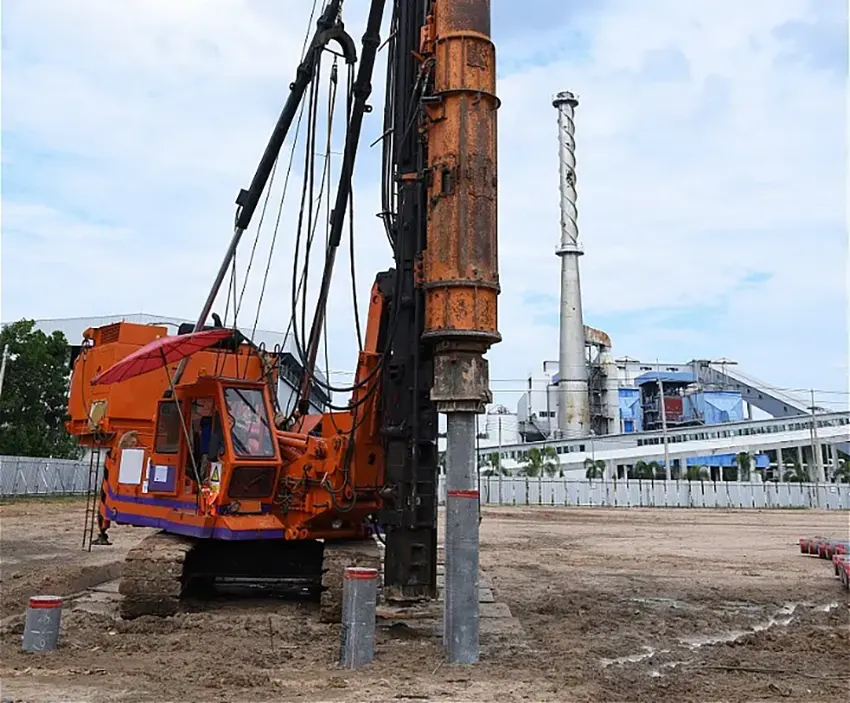 Heavy construction machine driving concrete piles into the ground at an industrial site with a factory in the background.