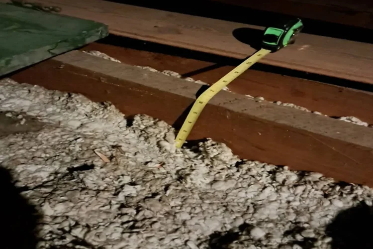 Cellulose insulation installation in progress in the attic of a property in Queens, NY with a measuring tape near it to measure the thickness installed.<br />
